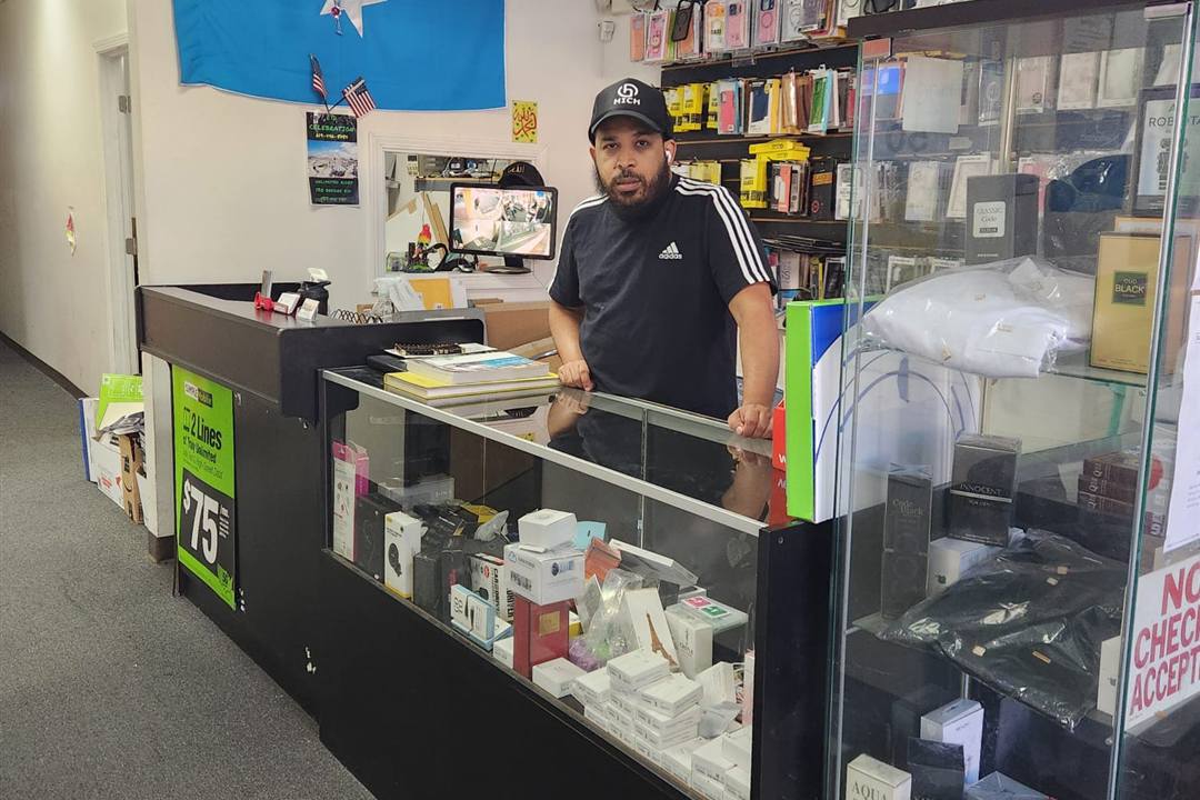 A Mobile City team member behind the glass counter, with phone cases and accessories on the wall behind him